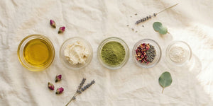 Various natural ingredients including oil, butter, green clay powder, and dried herbs on a light fabric background.